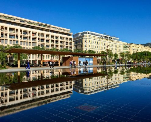 Vue urbaine ensoleillée avec des immeubles reflétés dans un miroir d’eau, des passants sur une promenade et un ciel bleu dégagé.